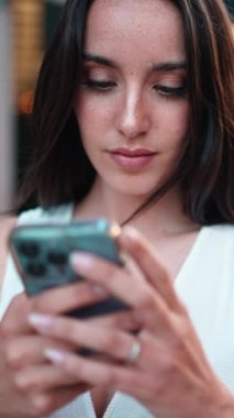 Clouse-up, çilli, koyu renk saçlı, beyaz bluzlu, elinde akıllı telefonuyla caddede dikilen kadın. Kız modern şehir arka planında cep telefonu kullanıyor