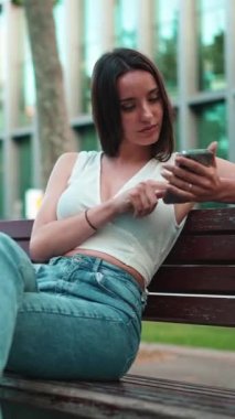 Çilleri ve koyu renk saçı olan güzel bir kadın elinde telefonla bankta oturuyor. Gülümseyen şirin kız fotoğraf izliyor, cep telefonuyla video çekiyor modern şehir arka planında.