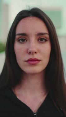 Çilli, açık kahverengi saçlı, siyah spor kapüşonlu genç bir kadın kameraya bakıyor ve modern şehir arka planının köprüsünde gülümsüyor.