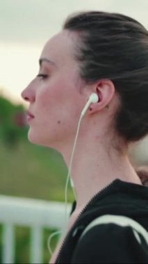 Çilli ve uzun at kuyruklu siyah spor kapüşonlu kulaklıklı genç bir kadın müzik dinliyor ve köprü boyunca yürüyor.