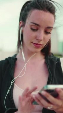 Çilli, uzun atkuyruklu, siyah kapüşonlu ve bej renkli spor bluzlu genç bir kadın cep telefonu kullanıyor.