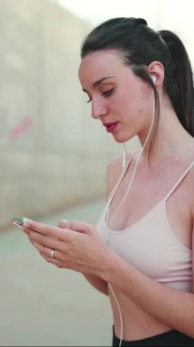 Uzun at kuyruklu, bej renkli kulaklıklı, elinde cep telefonu olan genç bir atletik kadın.