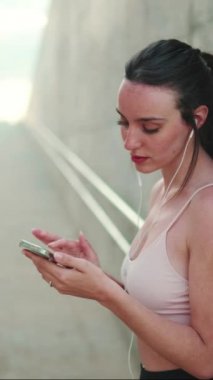 Uzun at kuyruklu, bej renkli kulaklıklı, elinde cep telefonu olan genç bir atletik kadın.