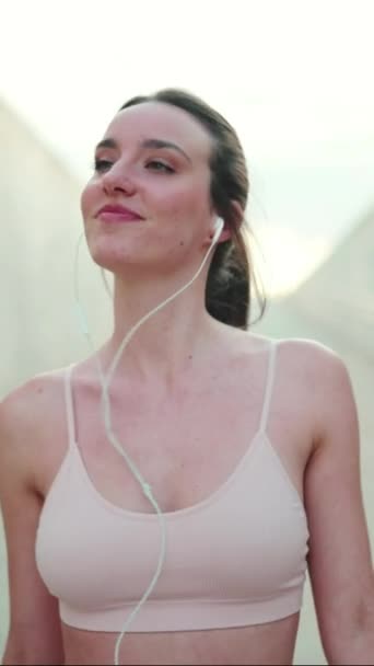 垂直ビデオ 配線されたヘッドフォンでベージュのスポーツトップを身に着けている長いポニーテールを持つ若い運動選手の女性 ウォーキングダンスと笑顔 — ストック動画