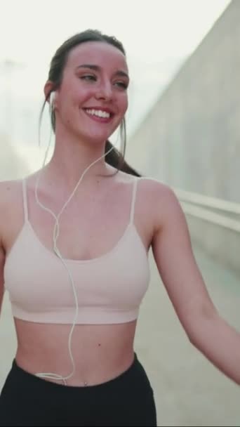 垂直ビデオ 配線されたヘッドフォンでベージュのスポーツトップを身に着けている長いポニーテールを持つ若い運動選手の女性 ウォーキングダンスと笑顔 — ストック動画