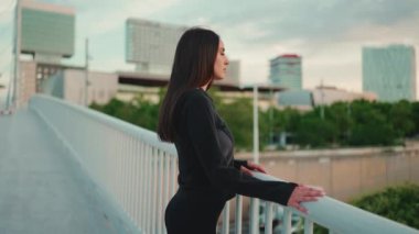 Çilli, açık kahverengi saçlı, siyah spor kapüşonlu güzel bir kadın köprüden modern şehre tepeden bakıyor. Kamera hareket ediyor kızım.