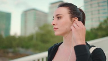 Çilli, uzun at kuyruklu, siyah kapüşonlu ve bej renkli spor bluzlu genç bir kadın kulaklarına kulaklık takıyor. Modern şehir arka planında köprüde dururken müzik dinliyor.