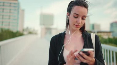 Çilli, uzun at kuyruklu, siyah kapüşonlu ve bej renkli spor bluzlu genç bir kadın köprü üzerinde dururken cep telefonu kullanıyor ve müzik dinliyor.