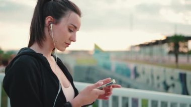 Çilli, uzun at kuyruklu, siyah kapüşonlu ve bej renkli spor bluzlu genç bir kadın köprü üzerinde dururken cep telefonu kullanıyor ve müzik dinliyor.