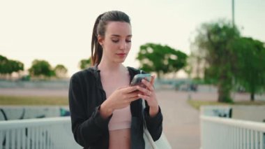 Çilli, uzun at kuyruklu, siyah kapüşonlu ve bej renkli spor bluzlu genç bir kadın modern şehir arkaplanının köprüsünde cep telefonu standı kullanıyor.