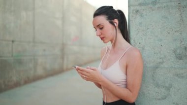 Uzun at kuyruklu, kablo kulaklıklı, bej rengi spor tişörtlü, elinde cep telefonu olan genç bir atletik kadın.