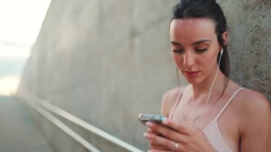 Uzun at kuyruklu, bej rengi kulaklık takan, elinde cep telefonu olan, takım elbiseli, atletik bir kadın.