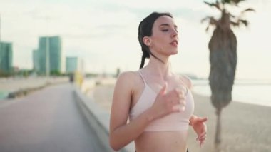 Örgülü atletik saçlı, bej rengi spor bluzlu, deniz arka planında kablosuz kulaklıklı genç bir kadın.