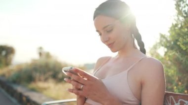 Örgülü atletik saçlı, bej renkli spor bluzlu genç bir kadın elinde cep telefonuyla bankta oturuyor, sosyal ağları, fotoğrafları ve videoları izliyor.