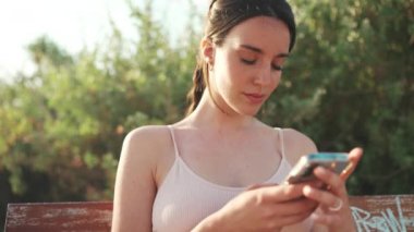 Örgülü atletik saçlı, bej renkli spor bluzlu genç bir kadın elinde cep telefonuyla bankta oturuyor, sosyal ağları, fotoğrafları ve videoları izliyor.