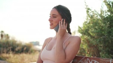 Saçı bej renkli spor bluzlu, akıllı telefondan konuşan sevimli, gülümseyen kız bankta oturuyor. Genç bir kadın cep telefonuyla iletişim kuruyor. Arka ışık
