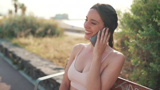 ベージュスポーツのトップを着てベンチに座ってスマートフォンで話をしているような笑顔の女の子 若い女性は携帯電話で通信する バックライト — ストック動画
