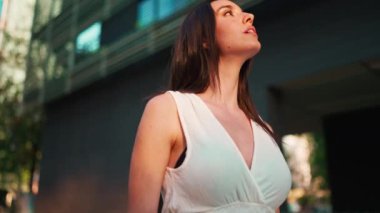 Çilli, koyu renk saçlı, beyaz bluzlu ve kot pantolonlu hoş bir bayan modern şehir manzarasının tadını çıkarıyor. Uzun kirpikleri olan güzel bir kız binanın üst katlarına bakıyor.