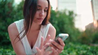 Çilli, siyah saçlı, beyaz bluzlu ve kot pantolonlu hoş bir genç kadın cep telefonu kullanıyor. Şehir parkında oturan güzel bir kız fotoğraflara, cep telefonundaki videolara bakıyor.