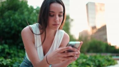 Çilli, siyah saçlı, beyaz bluzlu ve kot pantolonlu hoş bir genç kadın cep telefonu kullanıyor. Şehir parkında oturan güzel bir kız fotoğraflara, cep telefonundaki videolara bakıyor.