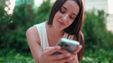 Çilli, siyah saçlı, beyaz bluzlu hoş bir bayan cep telefonu kullanıyor. Şehir parkında oturan güzel bir kız fotoğraflara ve cep telefonundaki videolara bakıyor. Kamera hareket ediyor