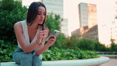Çilli, siyah saçlı, beyaz bluzlu hoş bir bayan cep telefonu kullanıyor. Şehir parkında oturan güzel bir kız fotoğraflara ve cep telefonundaki videolara bakıyor. Kamera hareket ediyor