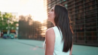 Çilli, koyu renk saçlı, beyaz bluzlu genç bir kadın sokakta dolaşıp etrafa bakıyor. Modern şehir geçmişinde güzel bir kız.