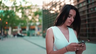 Çilli, siyah saçlı, beyaz bluzlu güzel bir kadın elinde akıllı telefonuyla sokakta yürüyor. Kız modern şehir arka planında cep telefonu kullanıyor