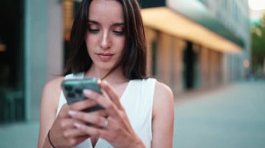 Çilli, koyu renk saçlı, beyaz bluzlu, elinde akıllı telefonuyla sokakta duran güzel bir kadın. Kız modern şehir arka planında cep telefonu kullanıyor