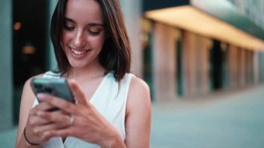 Çilli, koyu renk saçlı, beyaz bluzlu güzel bir kadın elinde akıllı telefonuyla sokakta duruyor ve gülümsüyor. Hoş kız modern şehir arka planında cep telefonu kullanıyor.
