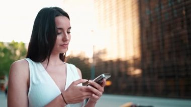 Çilli, koyu renk saçlı, beyaz bluzlu güzel bir kadın elinde akıllı telefonuyla sokakta duruyor ve gülümsüyor. Hoş kız modern şehir arka planında cep telefonu kullanıyor.