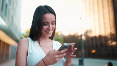 Çilli ve koyu renk saçlı güzel bir kadın ellerinde akıllı telefonları ve gülücükleriyle sokakta dikiliyor. Şirin kız modern şehir arka planında cep telefonu kullanıyor. Kamera hareket ediyor