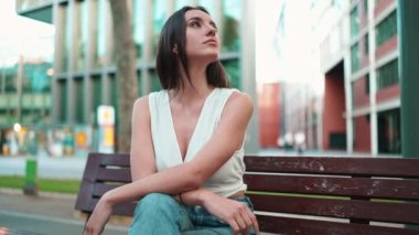 Çilli, siyah saçlı, beyaz bluzlu güzel bir kadın bankta oturur ve etrafına bakar. Şirin kız modern şehir geçmişinde oturuyor.