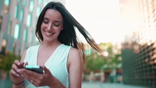 白いトップを身に着けているそばかすと暗い緩い髪を持つ美しい笑顔の女性は 彼女の手にスマートフォンで通りを歩いています 少女は現代都市の背景に携帯電話を使用します — ストック動画