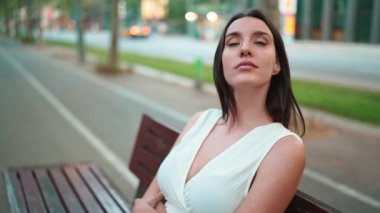 Çilli, siyah saçlı, beyaz bluzlu güzel bir kadın bankta oturur ve etrafına bakar. Şirin kız modern şehir geçmişinde oturuyor.