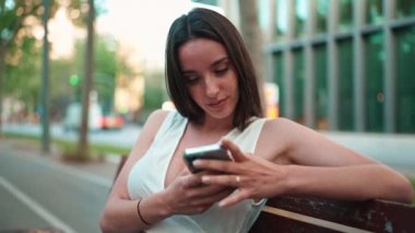 Çilli, koyu renk saçlı, beyaz bluzlu güzel bir kadın elinde telefonla bankta oturuyor. Gülümseyen şirin kız fotoğraf izliyor, cep telefonuyla video çekiyor modern şehir arka planında.