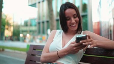 Çilli, siyah saçlı, beyaz bluzlu güzel bir kadın bankta oturuyor ve cep telefonundan mesajlar, fotoğraflar ve videolar izliyor. Kamera yaklaşıyor.