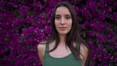 Çilli, koyu renk saçlı, yeşil bluzlu güzel bir kadın mor çiçeklerle süslenmiş bir duvara poz veriyor. Güzel kız hava öpücüğü gönderir ve kameraya bakarken gülümser.
