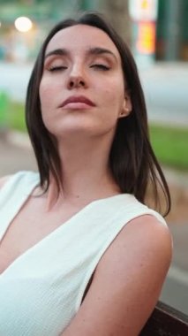 Çilli, koyu renk saçlı, beyaz bluzlu güzel bir kadın bankta oturur ve etrafına bakar. Şirin kız modern şehir geçmişinde oturuyor.