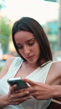 Çilleri ve koyu renk saçı olan güzel bir kadın elinde telefonla bankta oturuyor. Gülümseyen şirin kız fotoğraf izliyor, cep telefonuyla video çekiyor modern şehir arka planında.