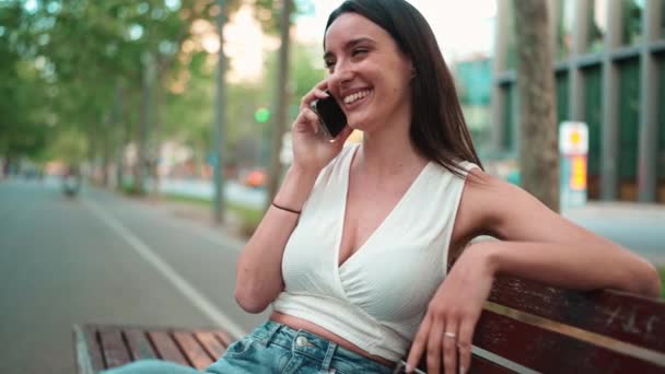 白いトップを身に着けているそばかすと暗い緩い髪の美しい女性は 彼女の手に電話でベンチに座っています Cute Girl Talking Mobile Phone Modern City Background — ストック動画