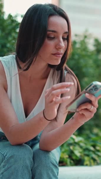 アダルトビデオ 白いトップを着た浅黒い髪の可愛い若い女性が携帯電話を使っています 美しい女の子は写真を通して見て都市公園に座っています 携帯電話上のビデオ — ストック動画
