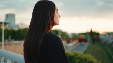 Çilli, açık kahverengi saçlı, siyah spor kapüşonlu bir kızın portresi kameraya bakıyor ve modern şehir arka planındaki köprüde yürüyor.