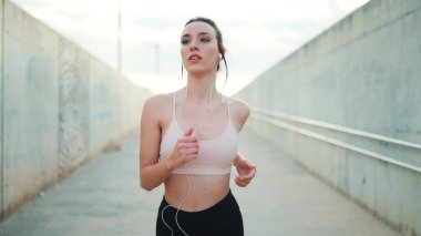 Uzun at kuyruklu, kablo kulaklıklı, bej rengi spor tişörtlü genç atletik kadın sahile doğru yaya geçidi boyunca koşuyor.