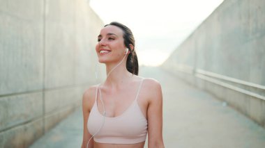 Uzun at kuyruklu genç atletik kadın. Telli kulaklıklarla bej rengi spor tişörtler giyiyor. Gülümsüyor.