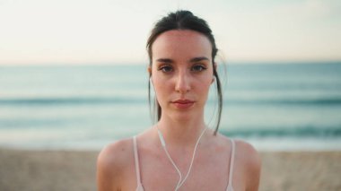 Uzun at kuyruklu genç bir bayan telli kulaklıklarla bej spor bir elbise giyiyor, deniz dibinde dururken kameraya gülümsüyor.