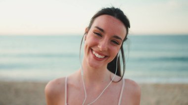 Uzun at kuyruklu genç bir bayan telli kulaklıklarla bej spor bir elbise giyiyor, deniz dibinde dururken kameraya gülümsüyor.