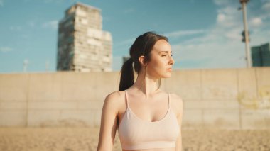 Uzun at kuyruklu, bej rengi spor bluzlu genç atletik kadın modern şehir arka planında şafak vakti kumsalda yürüyor.