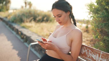 Örgülü atletik saçlı, bej renkli spor bluzlu genç bir kadın elinde cep telefonuyla bankta oturuyor, sosyal ağları, fotoğrafları ve videoları izliyor.