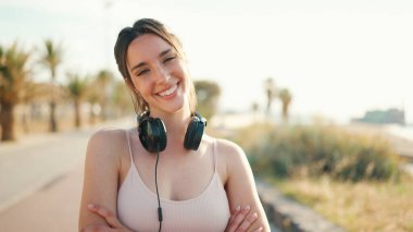 Saçları örgülü, bej renkli, kulaklık takan, setin üzerinde duran atletik bir kadın. Güzel kız elini çapraz yapar ve kameraya gülümser.